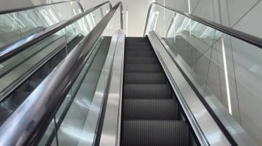 Modern yürüyen merdiven yukarı çıkıyor, yanında da aşağı doğru yürüyen bir yürüyen merdiven var. Parlak ışıklandırma parlak camlara ve metal yüzeylere yansıyor. Kentsel ulaşım kavramı