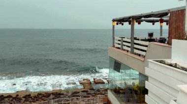Bulutlu bir günde okyanusa bakan bir deniz kenarı restoranı terası. Sıcak ışıklar ve asılı bitkiler rahat bir atmosfer yaratırken, kayalık kıyılara yumuşak dalgalar çarpar.