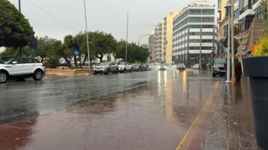 Şehirde yağmur yağıyordu. Arabalar parkın yanındaki ıslak yolda hareket ediyor, tekerleklerinden su fışkırtıyor. Binaların yansımaları su birikintilerinde görülebilir.