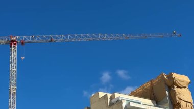 Açık mavi gökyüzüne karşı inşaat vinci. İki kurucu vinç bumbasına yerleştirildi ve yüksek mevkilerde çalışıyorlar. İşçiler koruyucu ekipman takıyor, kasklar ve emniyet kemerleri de dahil..