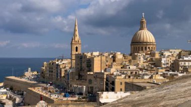 Valletta 'nın yüksek açılı manzarası: katedral kulesi, bazilika kubbesi, çatılar, arabalı yollar, sokaklardaki insanlar. Akdeniz ve arka planda fırtınalı bir gökyüzü.