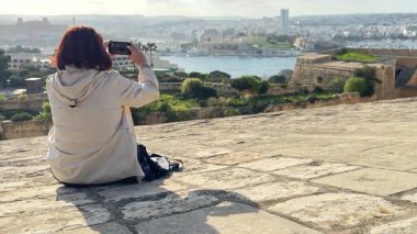 Taş yüzeyde oturan kadın turist, arkadan görülüyor, güneşli bir günde körfez ve tarihi kasabanın fotoğraflarını çekiyor..