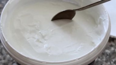 Açık bir kavanoz taze yoğurt yakın çekim. Metal kaşık, yoğun dokusunu ve pürüzsüz tutarlılığını vurgulamak için kremalı yoğurdu topluyor..