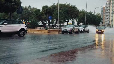 Üzerinde farları açık bir arabanın yüksek hızda sürdüğü yol işaretleri olan ıslak bir yol. Etrafta su uçuşuyor, asfalta büyük damlalar düşüyor. Arka planda çok katlı binalar, ağaçlı ve palmiyeli bir park, yağmurlu hava var..