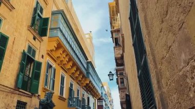 Geleneksel Malta sarı kireçtaşı mimarisi olan eski bir Avrupa kasabasında dar bir sokak. Bulutlu mavi gökyüzü, kuşlar, dekoratif elementli taş binalar, renkli ahşap balkonlar, yeşil pencereler ve panjurlar, bir sokak lambası