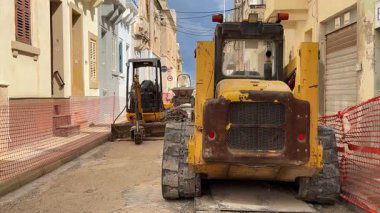 Yol çalışması sırasında dar bir caddede ağır inşaat malzemeleri. Bir kazıcı, yükleyici ve diğer makineler iş sahasında bir güvenlik ağı içerisindedir..