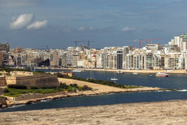 Ön planda tarihi kale ve arka planda modern binalarla deniz koyunun panoramik manzarası. Tekneler ve feribotlar derin mavi sularda ilerler beyaz bulutlu bir gökyüzünün altında.