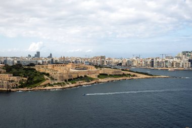 Ön planda tarihi kale ve arka planda modern binalarla deniz koyunun panoramik manzarası. Tekneler ve feribotlar derin mavi sularda ilerler beyaz bulutlu bir gökyüzünün altında.
