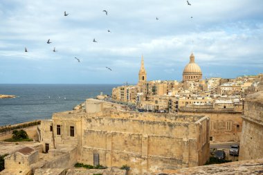 Valletta 'nın yukarıdan görünüşü: katedralin kubbesi, bazilikanın kubbesi, çatılar, arabalı yollar, sokaklardaki insanlar. Akdeniz ve arka planda mavi gökyüzündeki kuşlar..