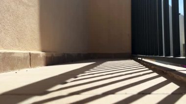 Metal bir çitin güçlü geometrik gölgeleriyle güneşli açık hava koridoru. Sıcak bej duvarlar ve fayanslı döşemeler minimalist ve modern bir estetik yaratıyor. Mimari ve soyut kavramlar için ideal.