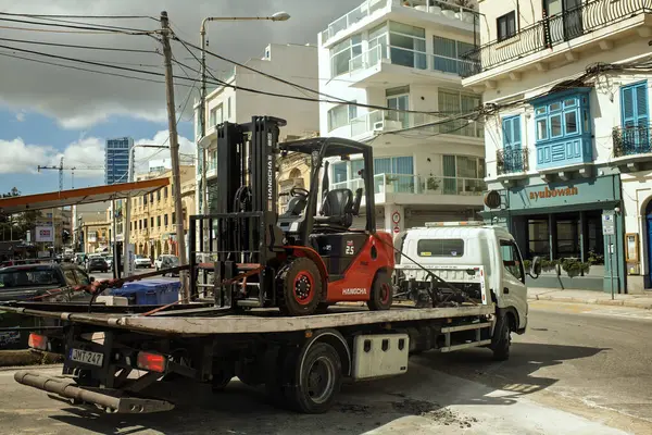 Gzira, Malta- 04.05.2025: Kırmızı forklift bir çekici kamyona yüklendi. Arka planda arabalar, benzin istasyonları, dükkanlar ve kafeler olan tipik hafta içi şehir hayatı.