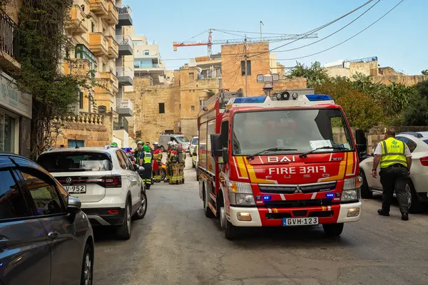 Gzira, Malta-04.05.2025: Mavi ışıklı modern kırmızı bir itfaiye aracı, konut binaları ve park halindeki arabalarla çevrili dar bir sokağa park edilmiştir. İtfaiye ve acil durum ekipleri çalışıyor. 