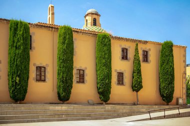 Parmaklıklı pencereleri olan eski sarı bir sıva duvarı boyunca simetri içinde duran uzun selvi ağaçları ve berrak mavi gökyüzünün altında renkli bir kubbe.