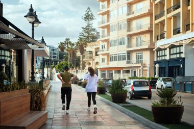 San Giljan, Malta - 04.08.2025: Palmiye ağaçları, modern binalar, kafeler ve arabalarla dolu bir cadde boyunca koşan genç erkek ve kadın. Sağlıklı yaşam tarzı kavramı. Editör kullanımı için