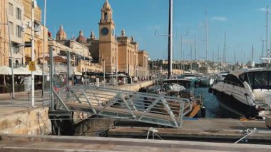 Malta 'daki huzurlu marina sahnesi: deniz kenarındaki kafeler, lüks yatlara giden bir iskele, açık mavi gökyüzü ve saat kulesi olan tarihi kireçtaşı binalar. Seyahat ve yaşam tarzı temaları için mükemmel.