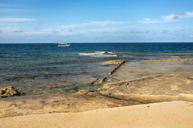 Kristal berraklığında mavi suları, kısmen batık doğal kaya oluşumları ve ufukta bir turist botu olan bir deniz manzarası. Akdeniz 'in güneşli havasıyla sakin bir sahil manzarası.