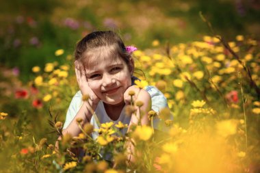 Sarı kır çiçekleriyle çevrili, çiçek açan bir çayırda oturan küçük bir kız kameraya gülümsüyor. Sahne masumiyet, neşe ve çocukluğun sıcaklığını yansıtıyor..