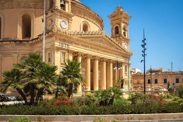 Mosta, Malta-05.21.2025: Büyük neoklasik sütunlar, bir heykel ve iki saat kulesinin yer aldığı Mosta Rotunda manzarası. Önde palmiye ağaçları ve renkli çiçekler, parlak mavi bir gökyüzüne karşı dizilmiş.
