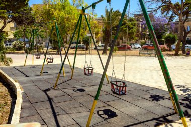 Güneşli bir günde şehir parkında, sessiz bir oyun parkında bir sıra boş çocuk sallanıyor. Gölgeler kauçuk paspasın üzerine düşer. Ağaçlar, arabalar ve binalar arka planda kalır. Huzurlu yaz havası.