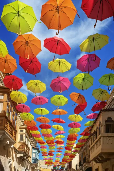 Parlak mavi gökyüzü altında geleneksel Akdeniz mimarisi olan bir caddenin üzerine asılmış parlak renkli şemsiyeler. Neşeli bir şehir yerleşimi şenlikli bir atmosfer yaratır..