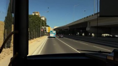 Marsa, Malta - 06.01.2025: Güneşli bir günde şehir caddesinde bir otobüsün ön görüntüsü, üstgeçit boyunca ilerliyor, metal bir köprünün altından geçiyor ve otobüs durağında duruyor..