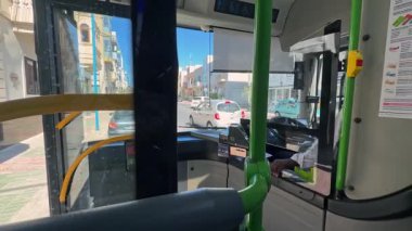 Fgura, Malta - 06.01.2025: Siyah tenli üniformalı sürücü belediye otobüsüne biner, direksiyona geçer, aynaları ve yolları dikkatle kontrol eder ve sürmeye başlar. Güneşli bir gün, ön koltuktan POV..