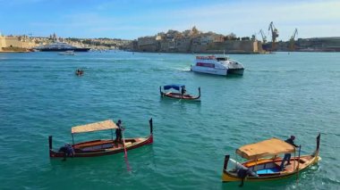 Valletta, Malta - 04.28.2025: Bir yolcu feribotu Maltas Grand Harbour 'dan geçiyor turkuaz sularda geleneksel Malta teknelerini geçiyor, Üç Şehir gökdelenleri ve tersaneleri görünüyor.