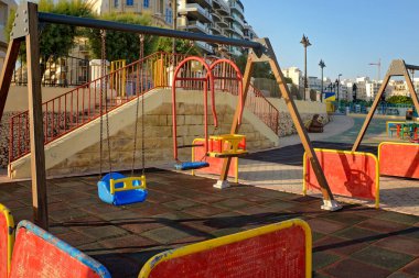 Gün batımında sakin bir şehir parkında boş renkli salıncaklar. Güvenli lastik döşeme, çitler ve arka planda modern binalar. Huzurlu bir açık hava sahnesi, çocuklar ve aile temaları için ideal..