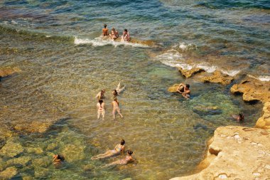 Sliema, Malta - 07.06.2025: Sığ ve temiz deniz suyunda doğal kaya oluşumlarıyla güneşli bir günün tadını çıkaran insanların hava görüntüsü.
