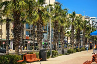 Gzira, Malta - 04.18.2025: Güneşli bir günde mağazaları, otelleri, kafeleri ve mavi gökyüzünün altındaki bankları olan işlek bir şehir yolu boyunca uzanan uzun palmiye ağaçları