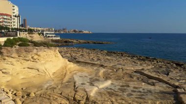 Akdeniz kıyısında Akdeniz kıyısında, Sliema, Malta, şehir gezinti alanı yakınında ve ufukta modern binalar, açık bir gökyüzü ve güneşli bir günde sakin bir deniz ile.