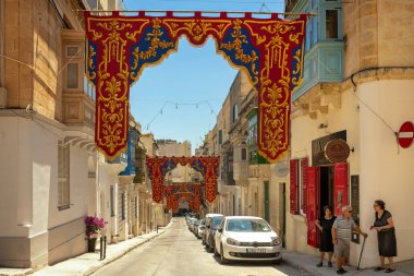 Sliema, Malta - 07.06.2025: Geleneksel balkonları, güzellik salonu ve park edilmiş arabaları ile sessiz dar bir cadde, Katolik Festa için renkli afişlerle süslenmiş.