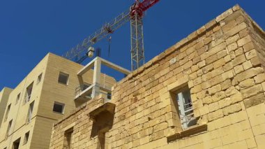 Kireçtaşı bloklardan yapılmış, pencere açıkları, beton bir çerçeve ve bitmemiş katlardan oluşan bir bina. Mavi gökyüzüne karşı bir vinç görünür..