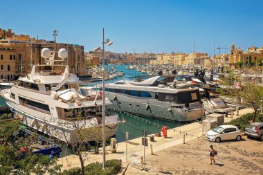 Birgu, Malta - 07.13.2025: Lüks yatları ve tekneleri olan güneşli bir liman, berrak mavi su boyunca demirli, parlak bir gökyüzüne ve kireçtaşı binalarla dolu bir şehir siluetine karşı..