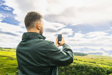 Yeşil ceketli genç beyaz adam Skye Adası 'ndaki İskoç İskoçya manzarasının akıllı telefonuyla fotoğraf çekiyor. Kopyalama alanı ile yatay olarak arka plan görüntüsü.