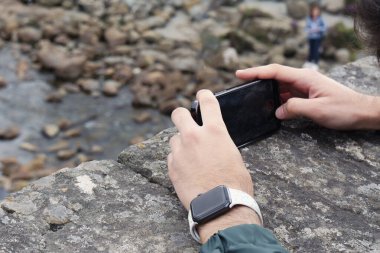 Sligachan, Cuilin Hills, İskoçya 'daki Eski Köprü' den akıllı telefonuyla fotoğraf çeken genç bir beyaz adamın elleri. Tanımlanamaz ve arka planda odak dışı bayan turist var. Yatay çekim.