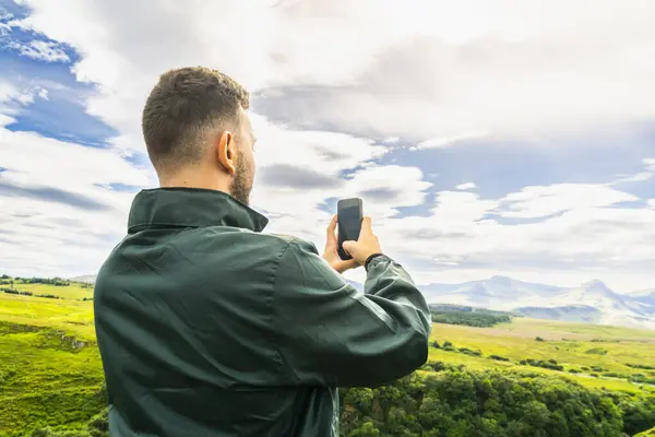 スカイ島のスコットランドのハイランドの風景のスマートフォンで写真を撮る緑のジャケットを着た若い白人男性 コピースペースで横に撮影したバックショット — ストック写真