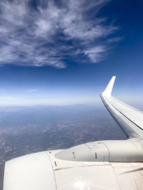 Uçak penceresinden bak. Mavi gökyüzünde ve aşağıdaki arazide duran beyaz bulutlar. Motorlardan biri ve ön planda bir kanat. Kopyalama alanı ile dikey çekim.