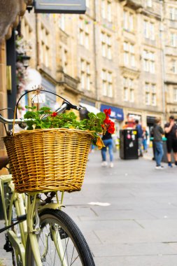 Edinburgh 'un Eski Kasabası' ndaki bir sokakta içi çiçek dolu bir bisiklet sepetinin yakın çekimi. Arka planda dikkati dağılmış, tanınmayacak bir yolcu var. Dikey çekim.