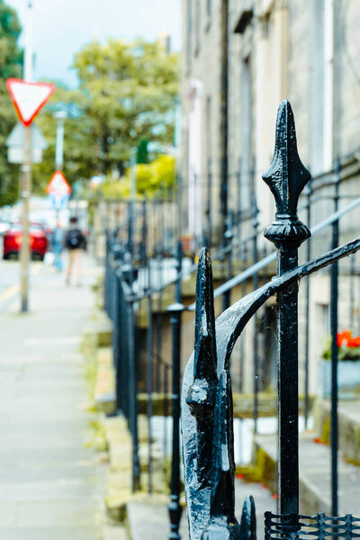 Detail of a street in downtown Edinburgh. Selective focus on a wrought iron gate in the shape of a spear. Traffic signs and passers-by out of focus in the background. Vertical shot.