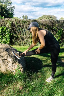 Genç İspanyol kadın parkta spor yapmadan önce esneme egzersizi yapıyor. Sağlıklı yaşam tarzı kavramları, yaz mevsimi bahar mevsimi. Kopyalama alanı ile dikey.