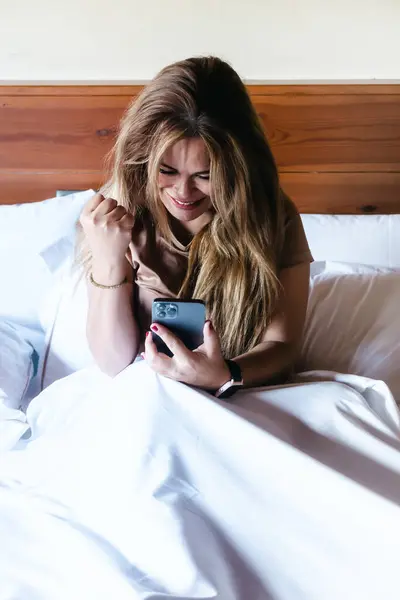 Young caucasian woman with smartphone in bed making winner and victory gesture. Vertical with copy space.