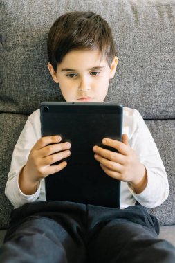 Evdeki gri koltukta oturan çocuk elektronik cihaz kullanıyor.