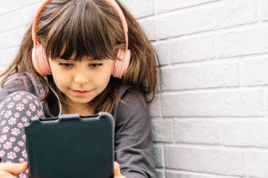 Dijital tablet kullanan ve kulaklık takan bir kız beyaz bir duvara yaslanmış.