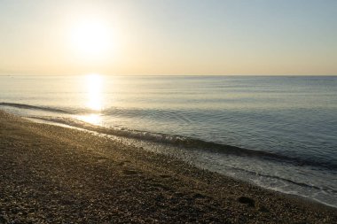 Sakin bir denizin üzerinde altın bir gündoğumu sırasında çakıl taşlı bir plajı yıkayan hafif dalgalar.