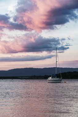 Durgun sulara demirlemiş beyaz yelkenli. Pembe ve mor günbatımı bulutlarını yansıtıyor. Dağları ve İskoçya 'daki köprüyü yansıtıyor.
