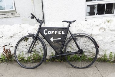 İskoçya 'da solunda ok ve kahve işareti olan beyaz bir duvara park edilmiş siyah antika bir bisiklet.