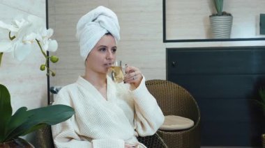 Kafasında havlu olan beyaz bornozlu bir kadın bir fincan meyve çayıyla spa salonunda oturur. Rahatlama, sağlık ve kişisel bakım kavramı
