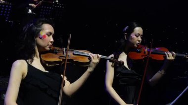 Etnik tarzda kemancı ikili dramatik sahne ışıkları altında gösteri yapıyor. Çiçekli siyah elbiseli iki Asyalı kemancı sahnede sanatsal ışıklandırma altında müzik çalıyorlar..