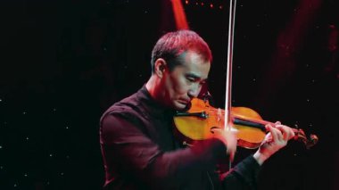 Profesyonel bir müzisyen, kırmızı ve mavi ışıklarla aydınlatılmış karanlık bir sahnede keman çalar..